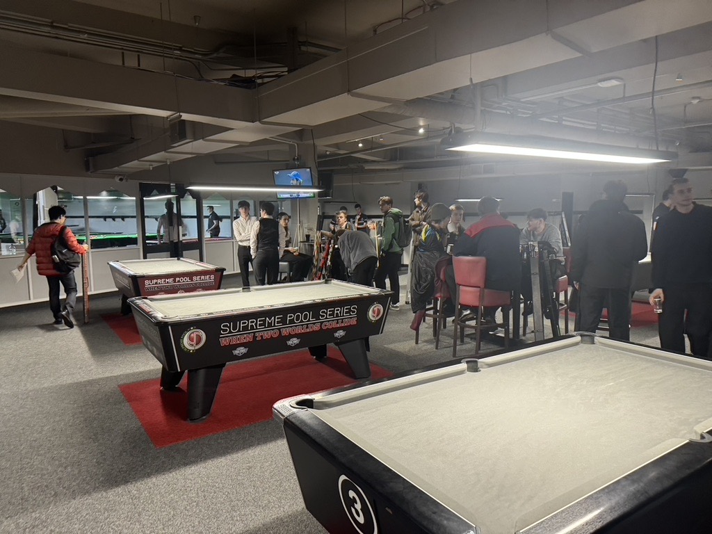 A university pool hall on a match night — Supreme Pool Series tables with players around them.