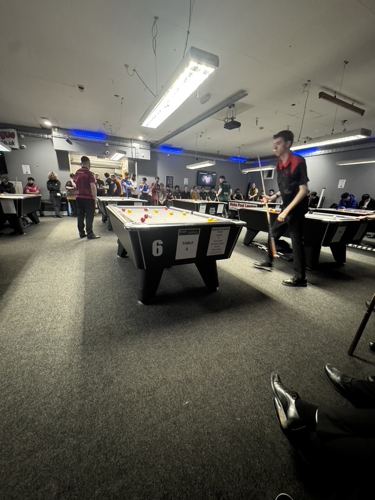 A player in a KCL pool shirt walking past table 6 at an 8-ball competition.